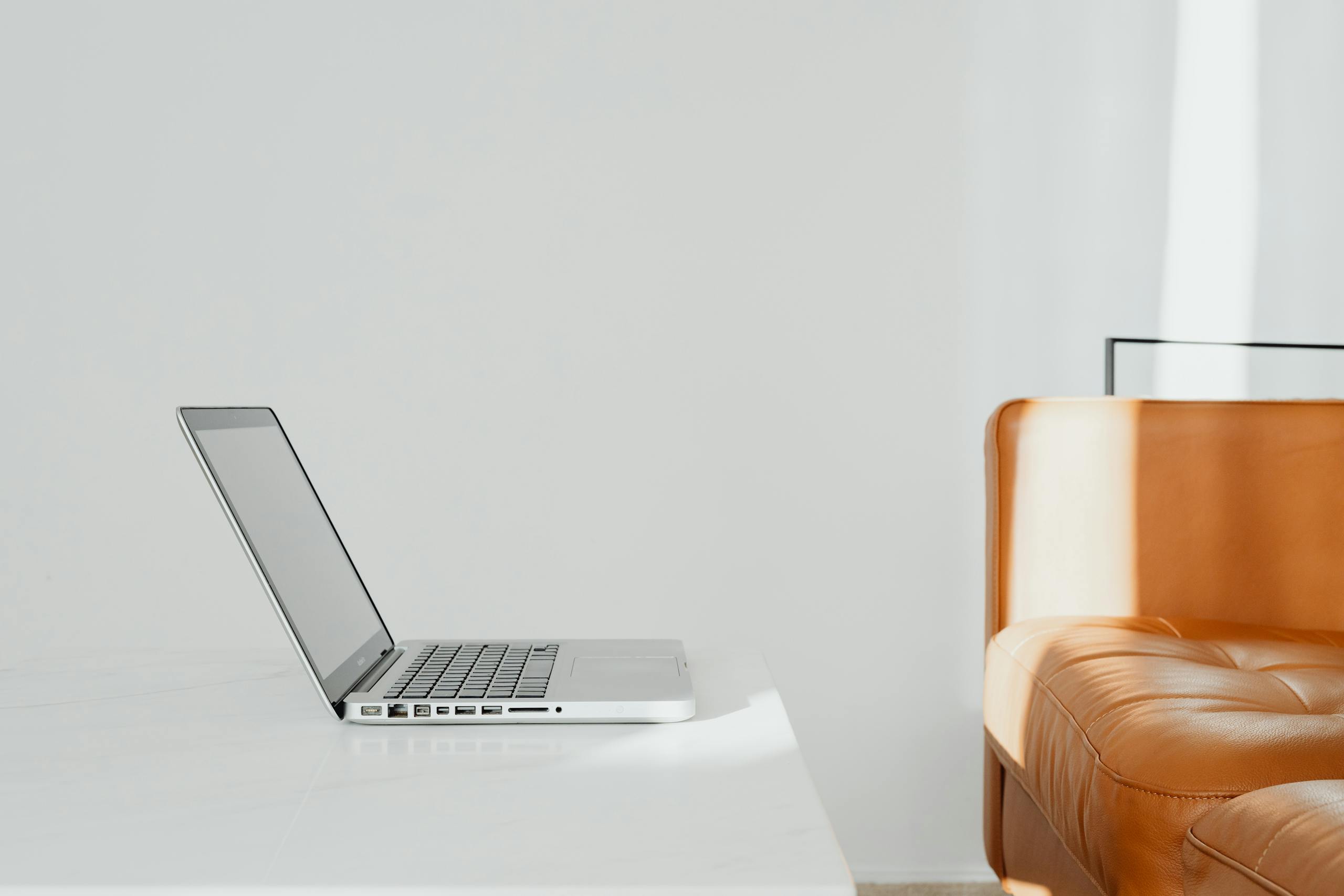 A bright and minimalist workspace with a laptop and orange leather sofa against a white wall.